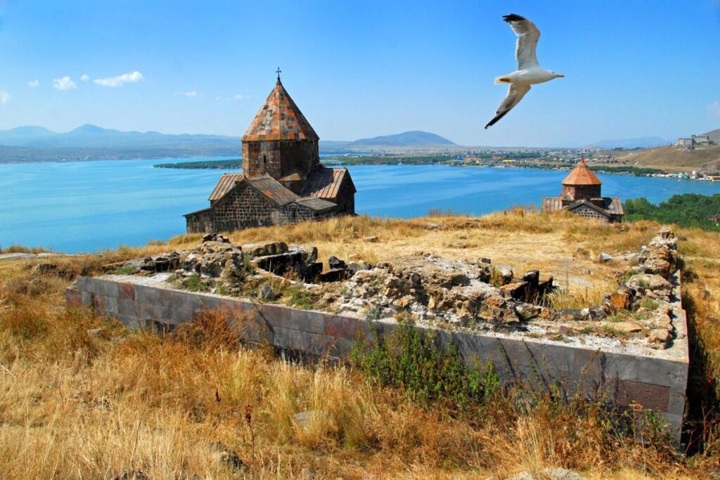 A picturesque view of Lake Sevan with the ancient Sevanavank Monastery on the shore and a seagull soaring in the clear blue sky.