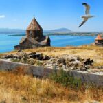A picturesque view of Lake Sevan with the ancient Sevanavank Monastery on the shore and a seagull soaring in the clear blue sky.