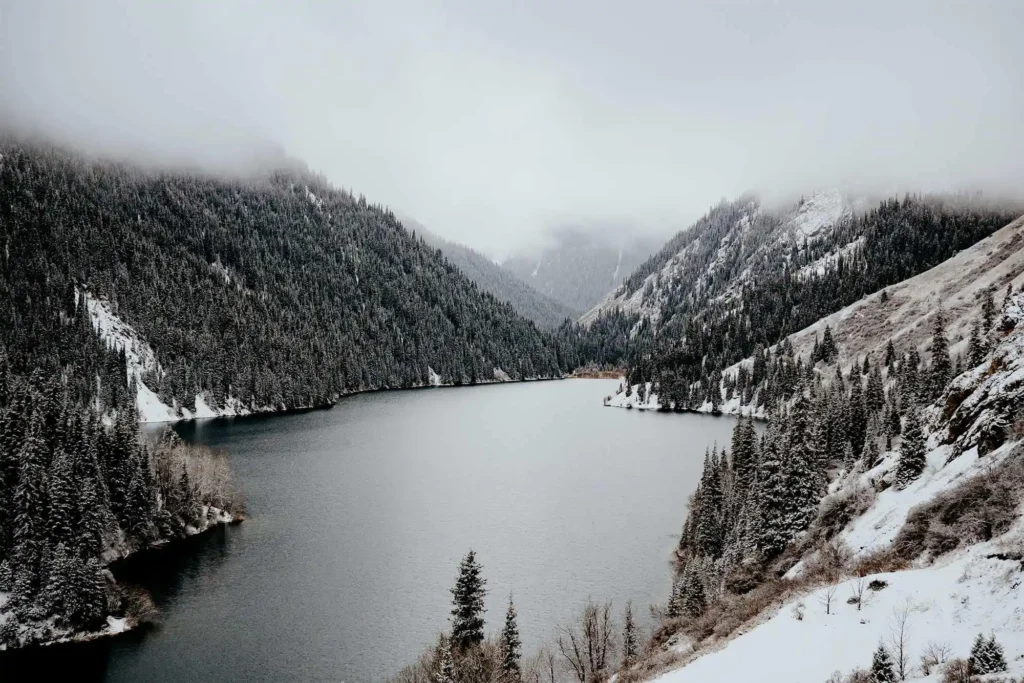 A peaceful winter scene of Kolsay Lake surrounded by snow-dusted pine trees and mist-covered mountains in Kazakhstan.