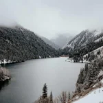 A peaceful winter scene of Kolsay Lake surrounded by snow-dusted pine trees and mist-covered mountains in Kazakhstan.