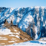 A scenic view of the Gergeti Trinity Church perched on a hill in Kazbegi, Georgia, surrounded by snow-covered mountains under a clear blue sky.
