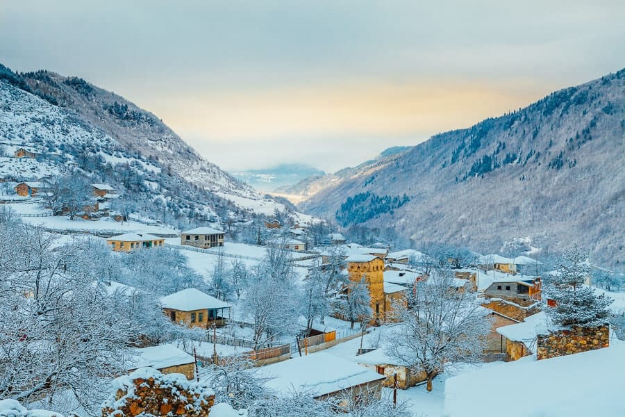 A picturesque snow-covered village nestled between the mountains in Georgia's Kazbegi region, surrounded by snowy landscapes and golden light.
