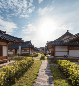 Hanok Village Traditional Korean hanok houses with tiled roofs in a peaceful village setting, surrounded by blossoming flowers and pathways under a bright sky.