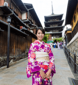 KIMONO IN GION A woman wearing a vibrant purple kimono with floral patterns, standing in a historic street in Gion, Kyoto, with traditional wooden buildings and a pagoda in the background.