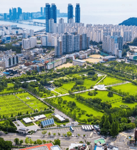 UN MEMORIAL BUSON An aerial view of the United Nations Memorial Cemetery in Busan, South Korea, with neatly arranged graves, manicured gardens, and the city skyline in the background.
