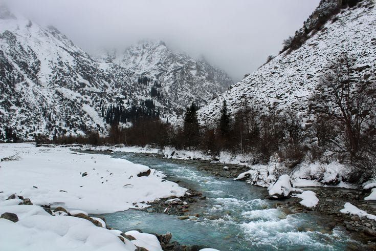 Snow-covered mountain landscape with a river flowing through Kyrgyzstan’s winter wilderness.