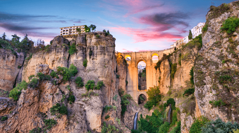 A breathtaking view of the Puente Nuevo bridge spanning a deep gorge in Ronda, Spain, surrounded by rugged cliffs and greenery under a colorful sunset sky.