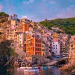 A vibrant sunset view of colorful cliffside houses in Riomaggiore, Cinque Terre, Italy, with boats docked along the calm sea.