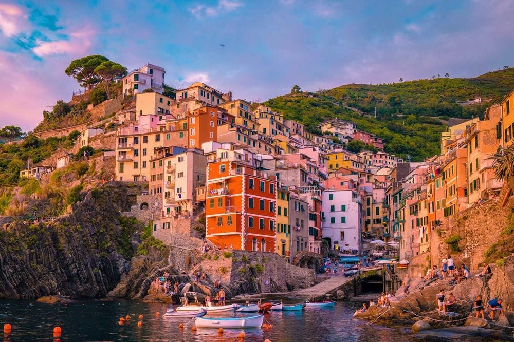 A vibrant sunset view of colorful cliffside houses in Riomaggiore, Cinque Terre, Italy, with boats docked along the calm sea.