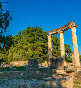 Olympia View of the ancient archaeological site of Olympia, Greece, showcasing the ruins of the Temple of Zeus, the ancient stadium, and surrounding columns, set amidst lush green hills.