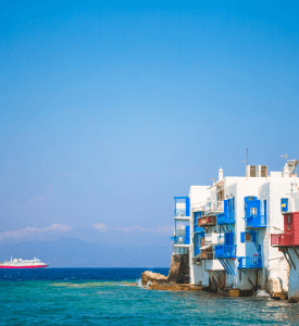 Mykonos Picturesque view of Mykonos, Greece, featuring iconic white-washed buildings, blue-domed churches, and vibrant bougainvillea flowers, with the crystal-clear waters of the Aegean Sea in the background.